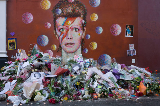 Portrait of David Bowie painted on a wall, with a pile of flowers and notes in front of it.