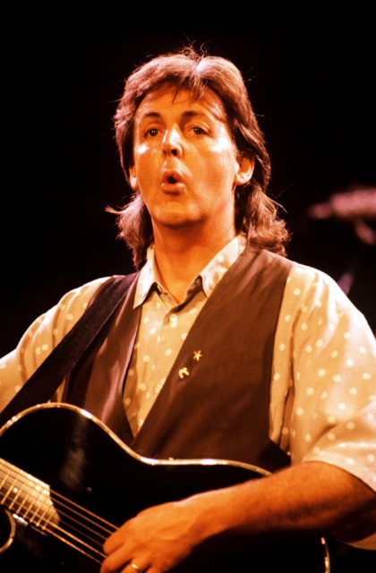 Paul McCartney wearing a vest, holding a guitar, and performing