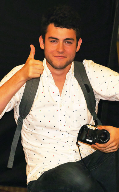 Truman Hanks in a white t-shirt with a backpack on holding a camera in one hand and giving a thumbs up with the other.