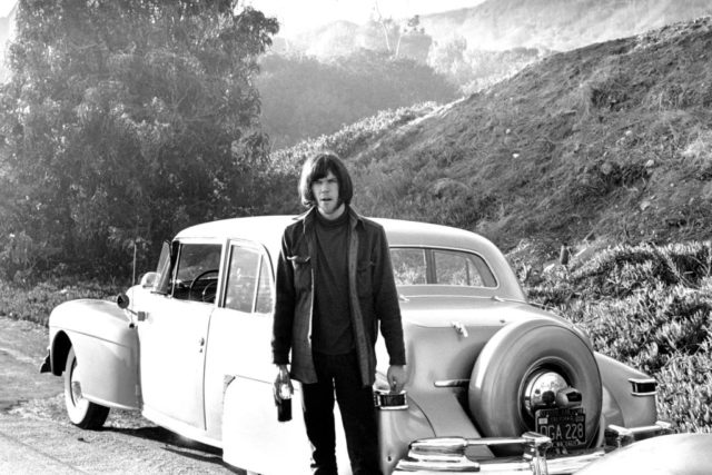 Neil Young standing in front of a car holding a glass bottle in one hand.