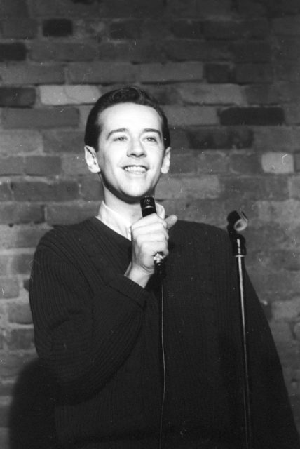 Headshot of a young George Carlin on stage holding a microphone