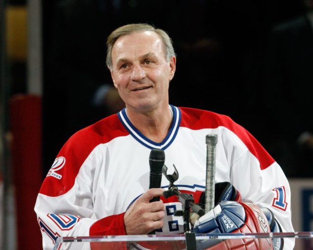 Guy Lafleur wearing a Montreal Canadians jersey and holding a microphone and hockey stick.