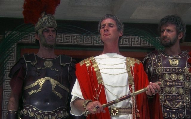 Michael Palin in a red and white roman robe, while John Cleese and Graham Chapman stand behind him dressed as gladiators. 