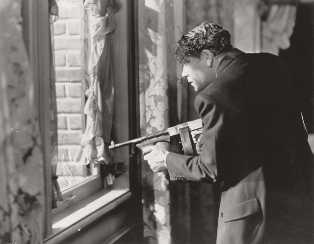 Paul Muni in a black jacket holding a gun that points out the window.