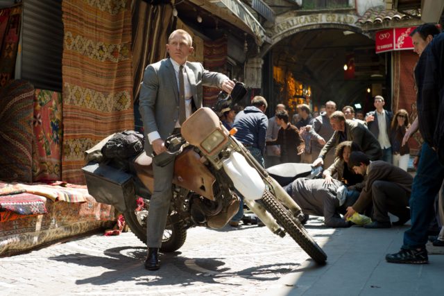 Daniel Craig as James Bond straddles a motorcycle in the middle of a busy street.