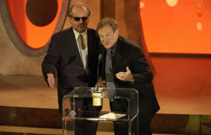 Jack Nicholson in a black suit and sunglasses stands beside Robin Williams, also in a black suit, behind a podium.