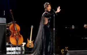 Naomi Ackie as Whitney Houston singing on stage in a long black dress with cape.