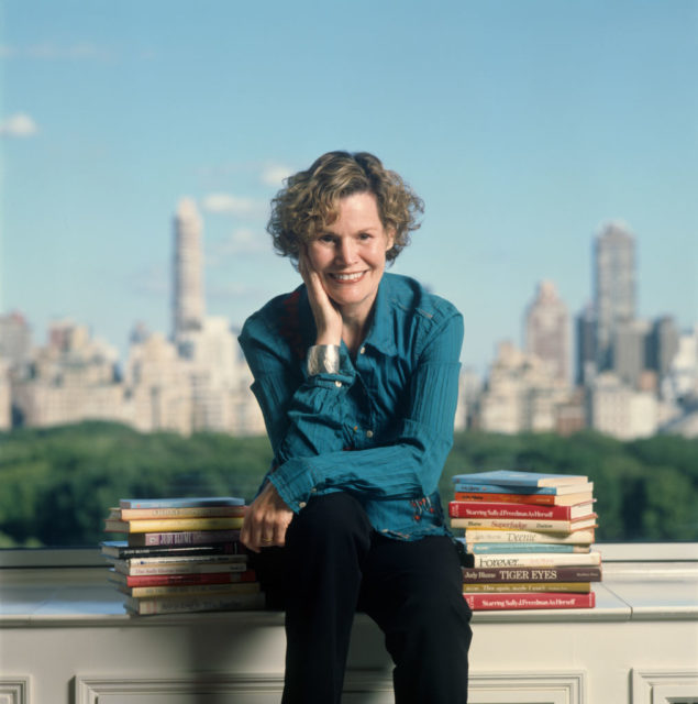 Judy Blume poses in front of a cityscape with piles of books beside her.