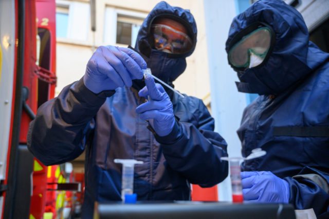 Two people in full cover blue suits with goggles and plastic gloves hold syringes. 