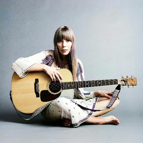 Joni Mitchell sits on the floor with a guitar