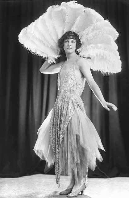 Flapper in a sparklingly dress holding up a large feathered fan behind her head.