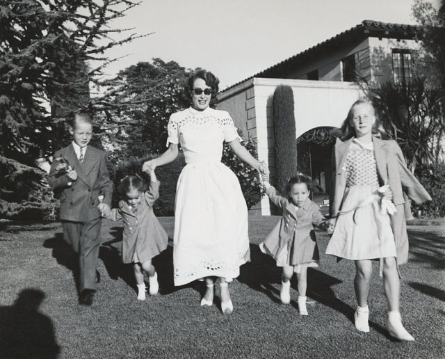 Joan Crawford in a white dress walking in a line holding hands with her four children.