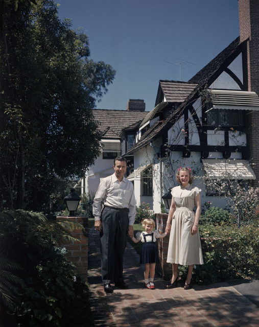 June Allyson and Dick Powell stand holding hands with their daughter Pamela in front of a house.