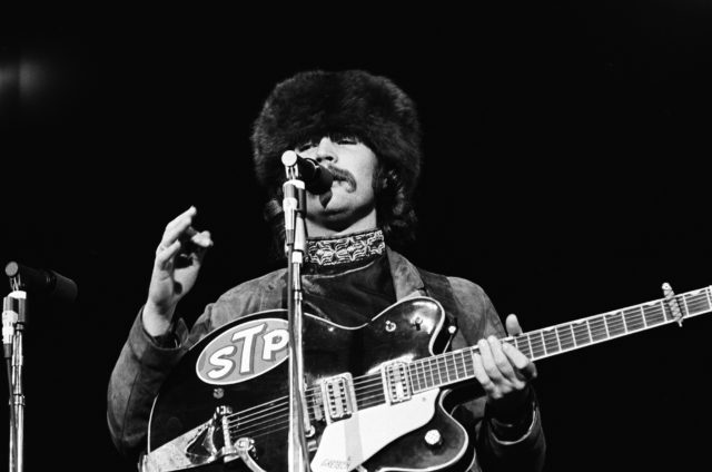 David Crosby in a fur hat, holding a guitar and speaking into a microphone.
