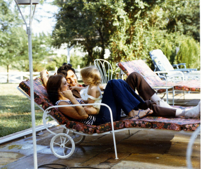 Priscilla and Elvis Presley lying on a sun chair with Lisa Marie sitting on their laps.