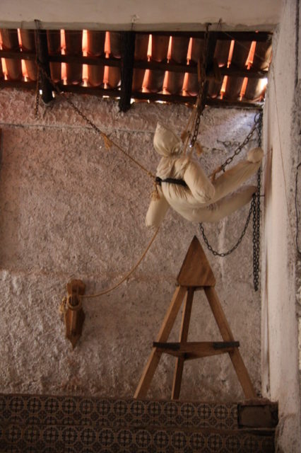 A dummy hangs by ropes and pulleys over a Judas Cradle in the corner of a stone room
