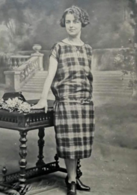Black and white photo of Lucile Ranon in a long plaid dress leaning against a table.
