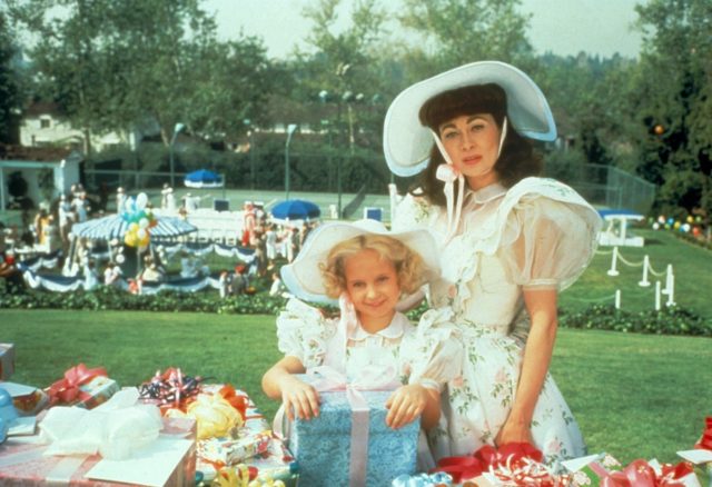 Faye Dunaway as Joan Crawford in a white dress with poofed sleeves and a large white hat, and Mara Hobel in a matching outfit.