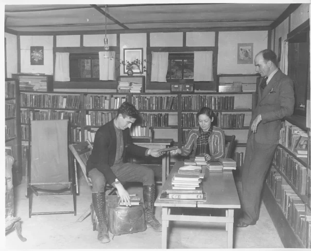 A woman and two men gathered in a library, sitting at a table full of books.