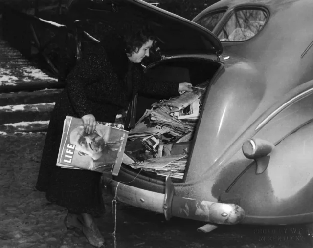 A woman pulling magazines from the trunk of an old car.