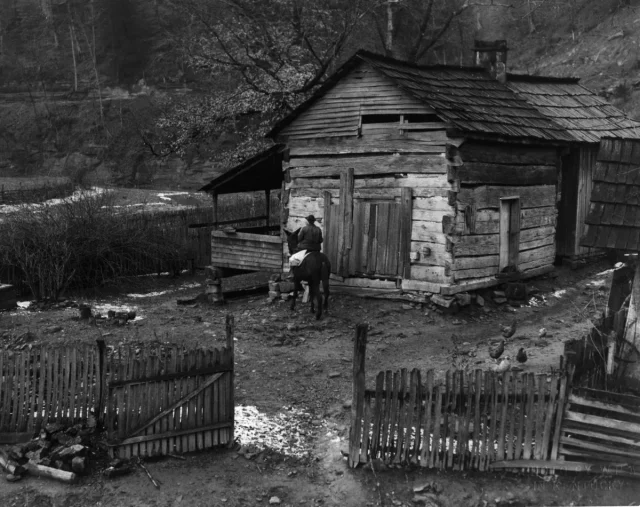 The back of a woman riding horseback towards a wooden cabin.