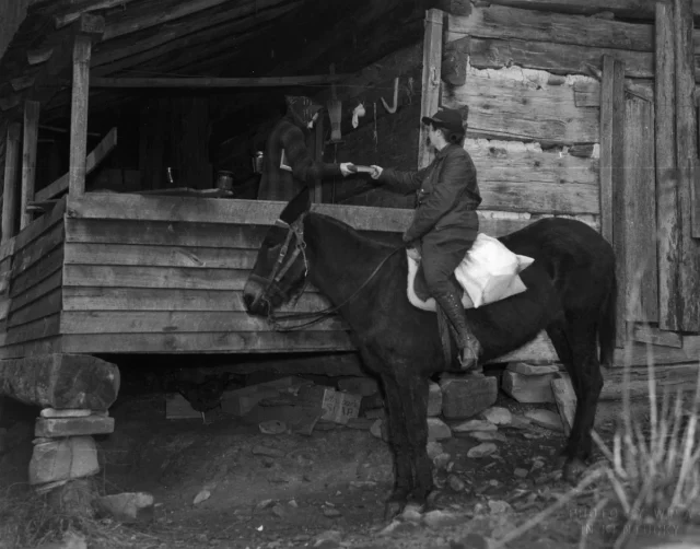 A woman on horseback handing another person a book at the side of their porch.