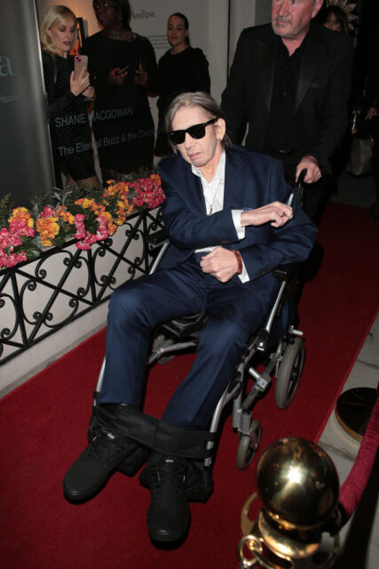 Shane MacGowan being pushed down a red carpet in a wheelchair