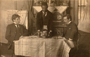 Two men sitting at a table being served another man's head on a plate