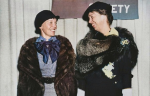 Amelia Earhart and First Lady Eleanor Roosevelt smile at each other while wearing elegant fur coats.