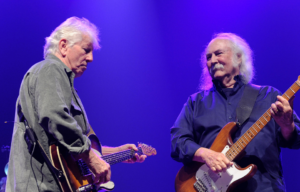 Graham Nash and David Crosby playing guitars on stage while facing one another.