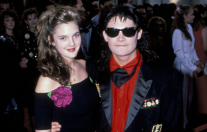 Drew Barrymore, wearing a black dress with roses, and Corey Feldman, wearing sunglasses and a red dress shirt, posing together for a photograph.