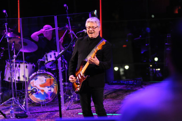 Steve Miller in all black sings and plays guitar on stage.