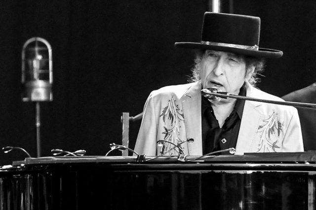 Bob Dylan in a black hat sits behind a microphone and a piano.