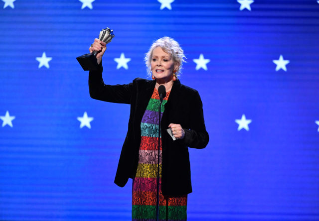 Jean Smart on stage, standing behind a microphone and holding up an award.