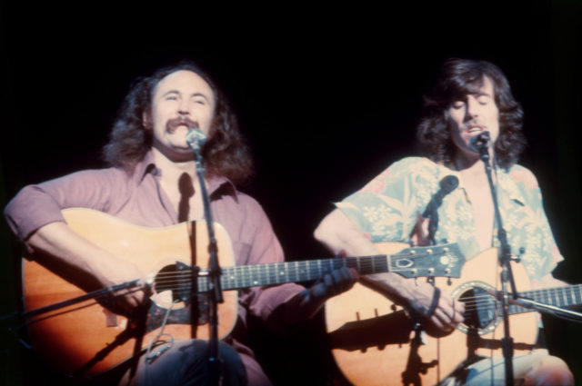 Young David Crosby and Graham Nash sitting on stage, playing guitars, and singing.