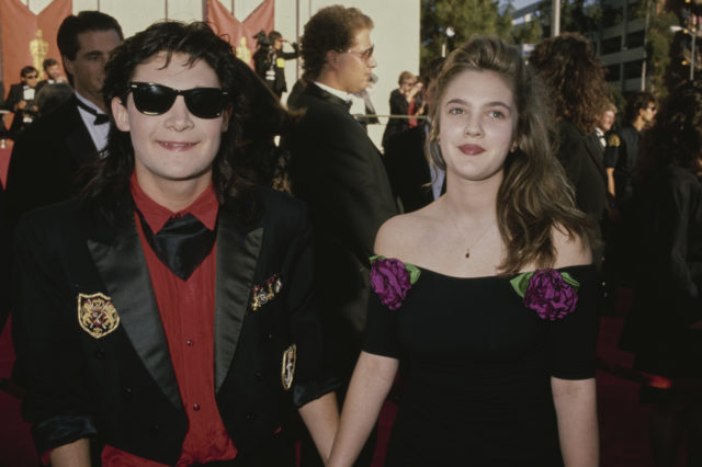 Corey Feldman and Drew Barrymore holding hands on the red carpet. Corey wears sunglasses and Drew wears a dress.