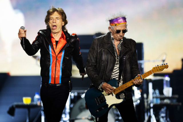 Mick Jagger in a blue and red jacket dances on stage beside Keith Richards playing the guitar in a leather jacket and zebra striped shirt.