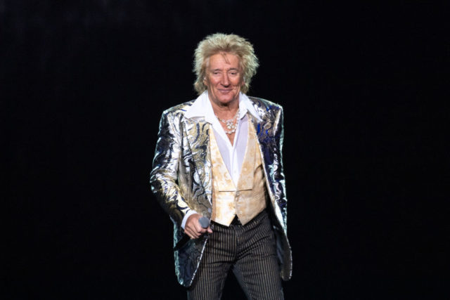 Rod Stewart in an elaborate shiny suit holds a microphone on stage and smiles.