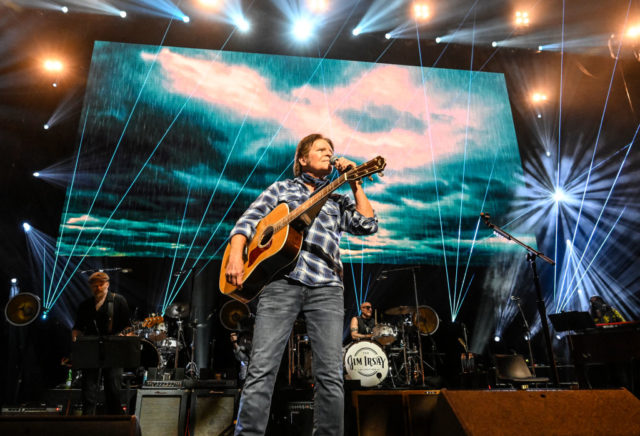 John Fogerty holds a guitar and a microphone on stage with a light show in the background. 