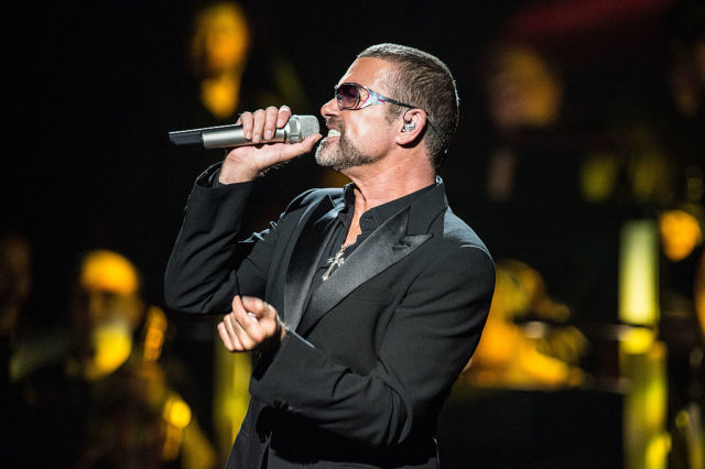 George Michael singing into a microphone while wearing an all black suit.