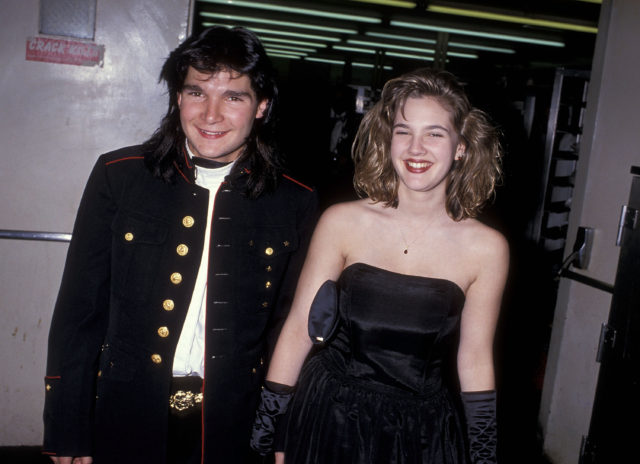 Corey Feldman and Drew Barrymore holding hands in a black suit jacket and a black dress.
