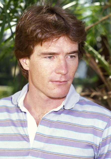 Headshot of a young Bryan Cranston in front of tree branches and wearing a striped polo shirt.