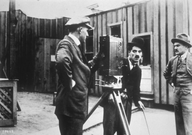 Charlie Chaplin behind a camera with two other men on set of a silent film.