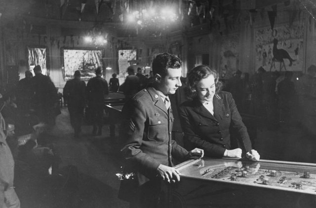 American soldiers play pinball in a busy officers club.