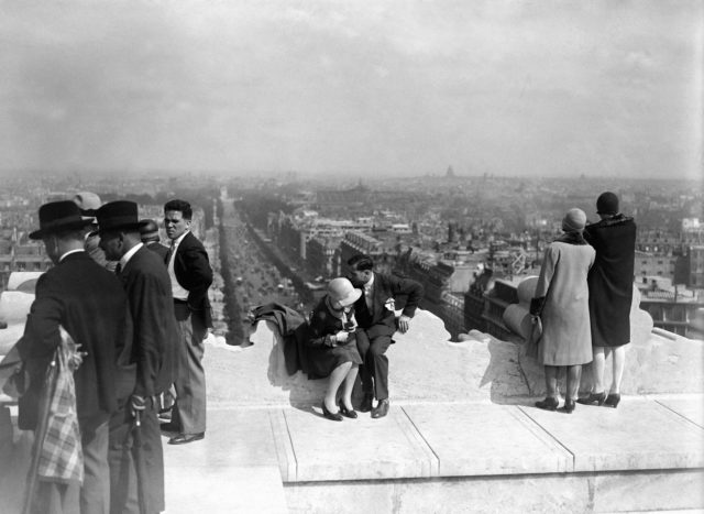 Tourists stand and sit on top of a roof overlooking a city.
