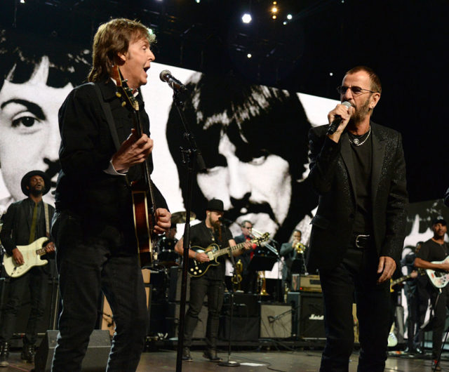 Paul McCartney holding a guitar sings into a microphone beside Ringo Starr holding a microphone, both in all black.