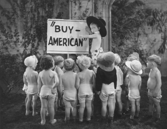 A young boy in a hat points to a sign that says "Buy American" while a group of other toddlers in diapers surround him.