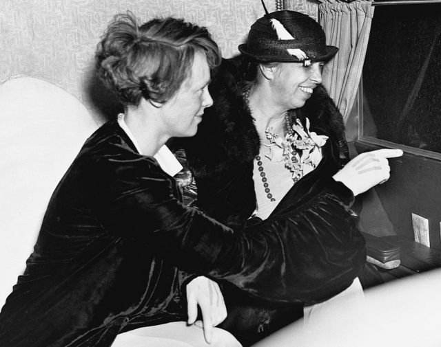 Amelia Earhart pointing something out an airplane window to First Lady Eleanor Roosevelt who sits beside her.