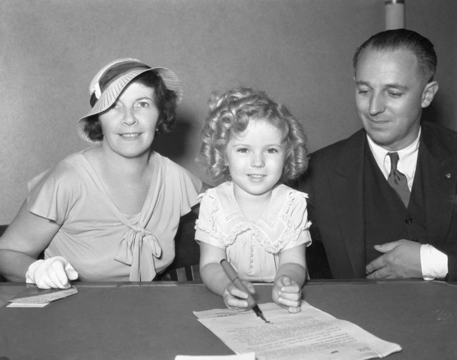 Shirley Temple signing a paper with her parents.