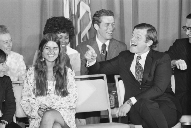 Edward Kennedy sitting and pointing to the side with other people sitting around him. A young Maria Shriver is laughing. 